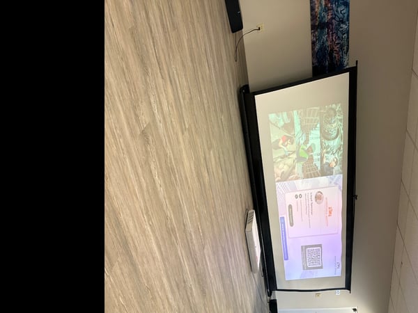 A rotated photograph capturing the corner of an indoor room featuring light wood paneling and a projection screen displaying a presentation slide. The slide shows industrial images, a profile picture, contact details for 'Lloyd Spencer,' and a QR code.