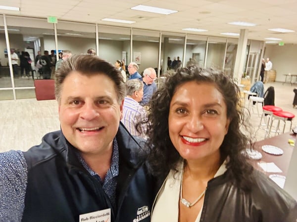 A close-up selfie of a smiling man and woman taken indoors at a social or networking event, with many other attendees visible socializing in the brightly lit background.