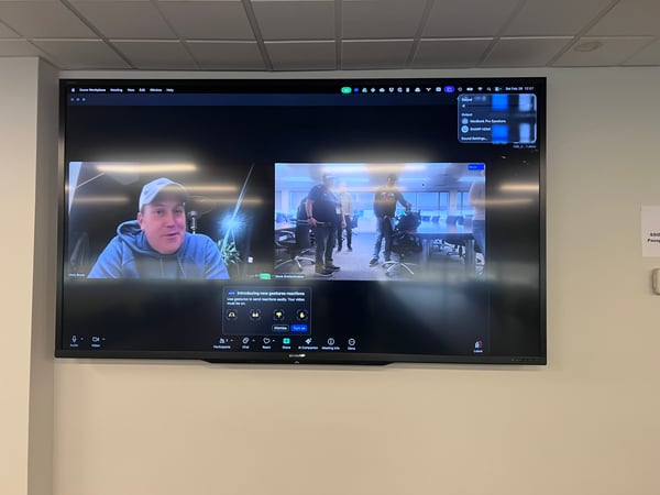 A large wall-mounted monitor displays a Zoom video conference call with two active video feeds and various interface overlays.
