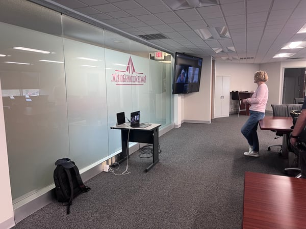 An indoor office environment featuring a woman standing near a desk, a glass partition with a mirrored logo, and a large wall-mounted screen displaying a video call.