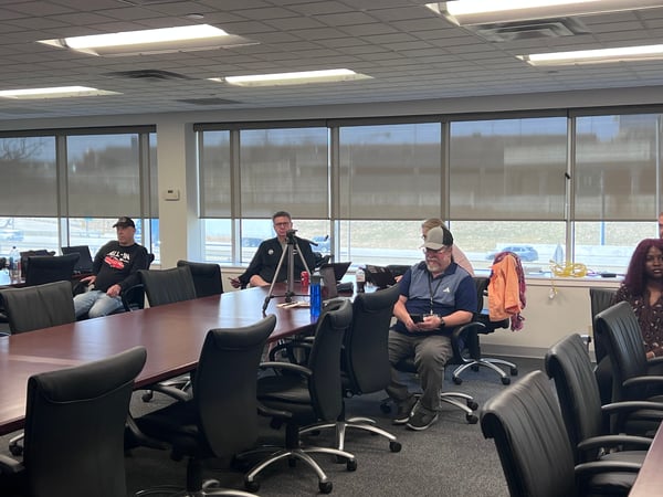 A group of people are seated around a large conference table in a modern office or meeting room, some looking at their phones or laptops.