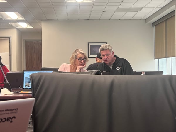 A man and a woman are seated at a desk in an office or classroom environment, intently looking at a laptop screen together.