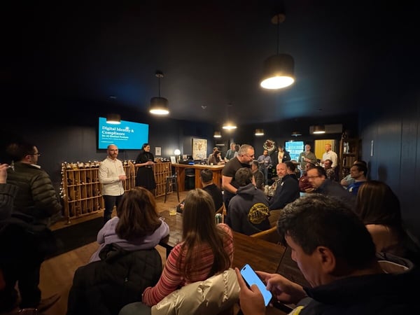 A low-light photograph capturing an indoor presentation or networking event where two speakers stand in front of a screen displaying the title 'Digital Identity & Compliance' to a large audience seated and standing in a dark, cozy venue.