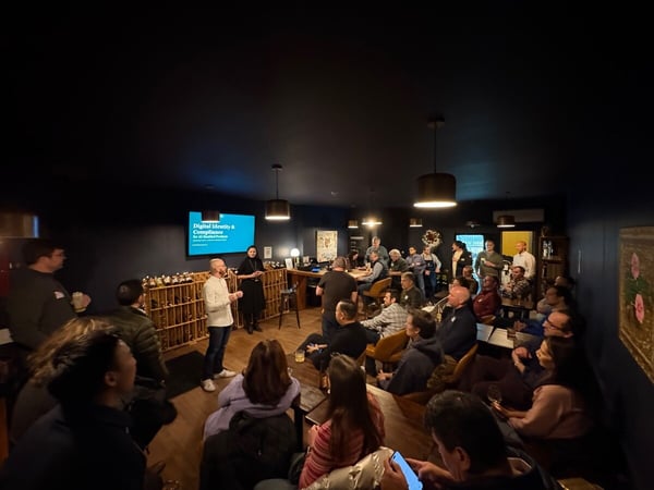 A candid photo captures an indoor professional gathering or presentation in a dark room, where a speaker stands next to a screen displaying "Digital Identity & Compliance" while addressing a crowded audience.