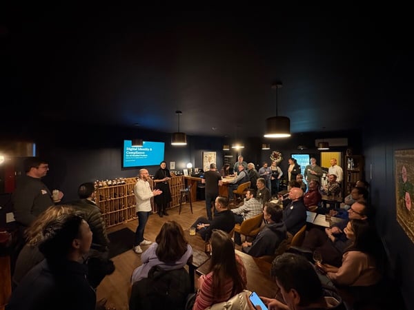 A photograph captures a technical presentation or meetup taking place in a dimly lit, intimate indoor venue with dark blue walls, showing a speaker addressing a large audience gathered around tables.