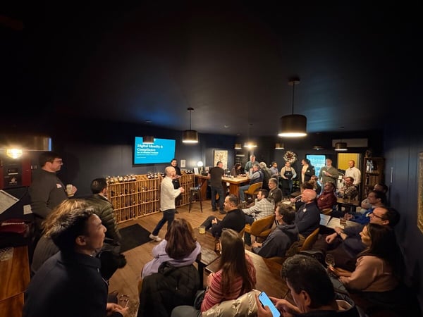 A candid photo capturing an indoor presentation or networking event in a dimly lit, dark-walled room, where a large group of attendees is seated and standing, listening to a speaker near a screen displaying a title slide.