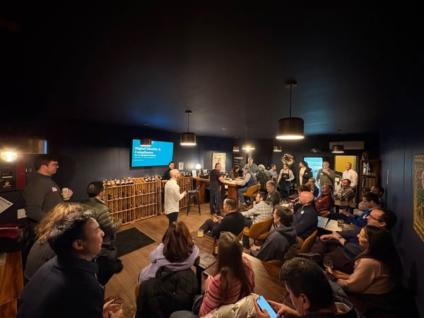 A low-light photograph capturing an indoor event or presentation where a speaker in a white shirt addresses a large, mixed audience gathered in a dark-walled venue that features a wine rack.