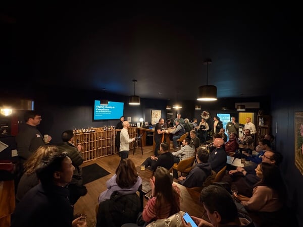A photograph captures a professional or networking event taking place indoors in a dimly lit venue with dark walls, where a speaker addresses a large seated audience gathered around tables and chairs.