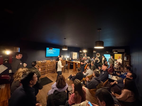 A dimly lit indoor photograph capturing a professional networking or educational event where a speaker is presenting to a large audience gathered in what appears to be a lounge or wine bar setting, with a presentation slide visible on a screen.