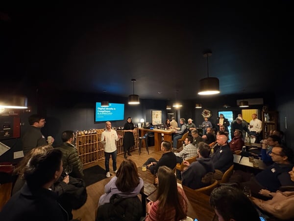A photograph capturing an indoor presentation or talk in a dimly lit venue, where two presenters stand in front of a screen addressing a large audience seated in chairs and along a bar.