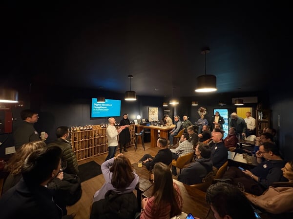 A low-light candid photograph captures an indoor professional or educational event taking place in a dark lounge, showing two presenters standing near a screen while addressing a large audience seated in rows of chairs and benches.