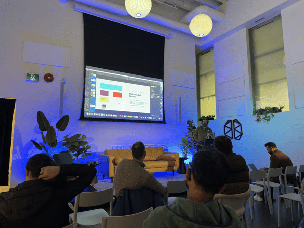 A photograph capturing an indoor technology presentation or meetup, where a male presenter stands near a podium addressing an audience seated in chairs, while a large screen displays a slide titled "Multimodal Models."
