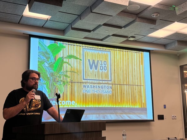 A man wearing glasses and a black t-shirt is giving a presentation, standing at a dark podium and speaking into a microphone in an indoor venue with distinctive acoustic ceiling tiles. Behind him, a projection screen displays a slide featuring a large 'W 1000' logo, 'WASHINGTON ONE THOUSAND', and the word 'welcome.'