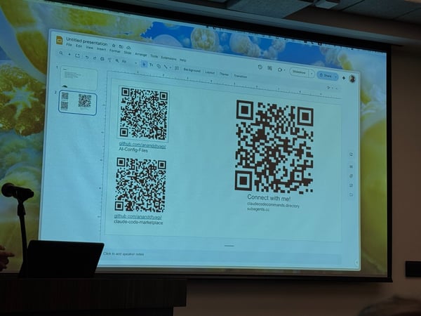 A photograph taken in a lecture setting showing a large projected screen displaying a presentation slide that features three QR codes and associated links, encouraging connection with the presenter. A microphone stand and a laptop are visible on a podium in the foreground.