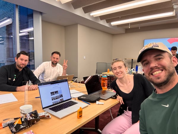 A group of four people, smiling and gathered around a table with open laptops and snacks, pose for a selfie in a modern meeting room during the evening.