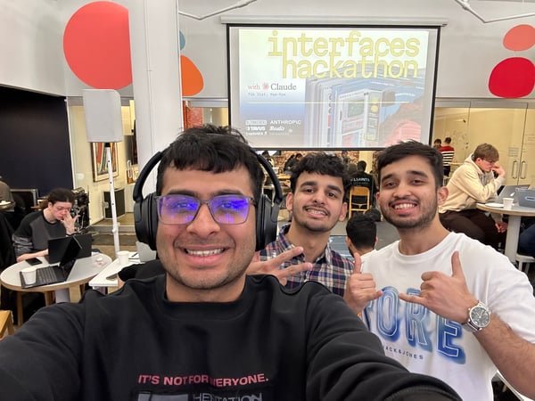 A group of three young men taking a selfie at an indoor event called 'interfaces hackathon with Claude'. The background shows other participants working at tables and a projector screen displaying event details.