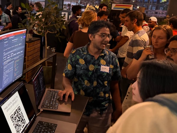 A candid photograph taken at a crowded indoor tech meetup or hackathon, showing attendees gathered around a table where two men are demonstrating code on a curved monitor and a laptop displaying a QR code prompt.