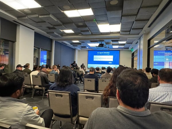 A photograph showing a large audience seated in a modern conference room, looking toward a projection screen that displays a system message indicating "No signal detected."