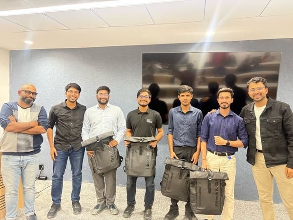A group portrait of seven men standing side-by-side indoors, likely in a modern office or conference room, with several of them holding dark gray backpacks.