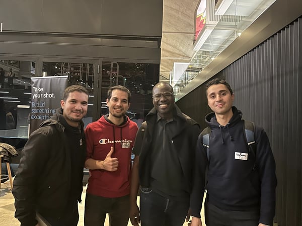 A group photo of four men smiling and posing together indoors, likely at an event or conference, with a motivational banner visible in the background.