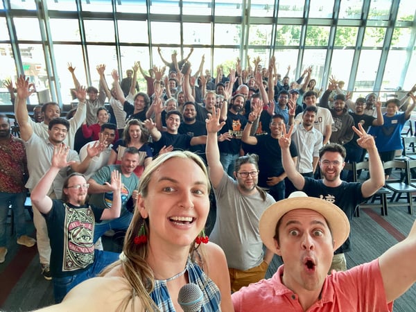A large group of approximately fifty to sixty people poses enthusiastically for a group selfie indoors, raising their hands and making gestures like peace signs in a bright conference room with large window walls.