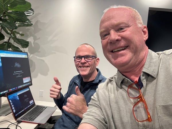 A close-up photograph of two smiling men giving thumbs-up gestures while seated at a desk with a laptop and an external monitor, which displays a cloud-related error message.
