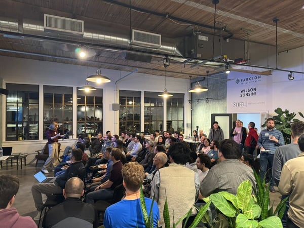 A candid photograph captures a large indoor event where a woman is giving a presentation to a seated audience in a loft-style venue with industrial architectural elements. Sponsor logos are visible on the back wall.