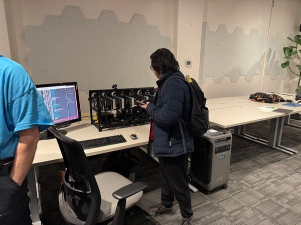 A photograph taken in a modern office space showing two individuals examining a specialized computer rig (likely a GPU setup) next to a monitor displaying technical code or log files.