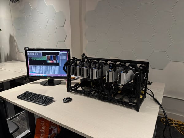 A photograph of a workspace featuring a desktop computer setup next to a large, open-frame rig containing multiple graphics processing units (GPUs), likely used for cryptocurrency mining or high-performance computing. The wall behind the desk is decorated with light gray hexagonal acoustic panels.