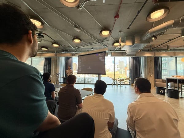 A group of people are seated in a modern office space, watching a presentation displayed on a projection screen with a city view in the background.
