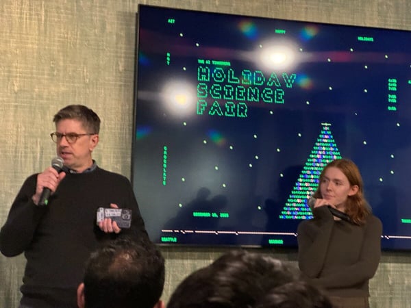 A photograph showing two presenters, a man and a woman, standing in front of a large digital screen at an indoor event, likely a science fair or holiday gathering.