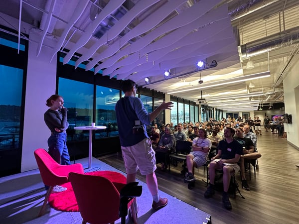 A photograph captures a speaker presenting to a large seated audience in a modern, well-lit office or event space at dusk. The room features distinctive wave-like ceiling panels, large windows overlooking a waterfront, and atmospheric purple and blue stage lighting.