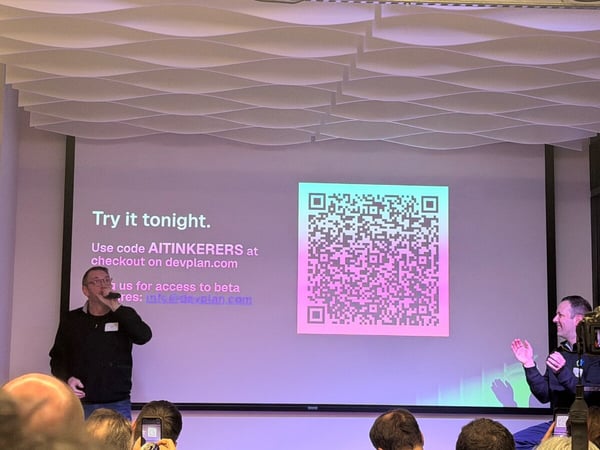 A man is giving a presentation in front of a large screen displaying a QR code and promotional text, while an audience watches from the foreground.