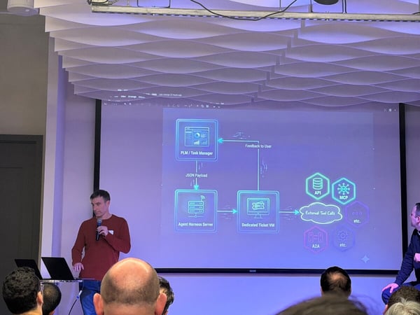 A man stands on a stage giving a technical presentation in front of a screen displaying a system architecture diagram.
