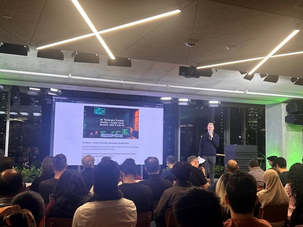 A photograph capturing an indoor tech meetup or presentation where a speaker addresses a large, seated audience in front of a large screen displaying event details for "AI Tinkerer's Toronto."