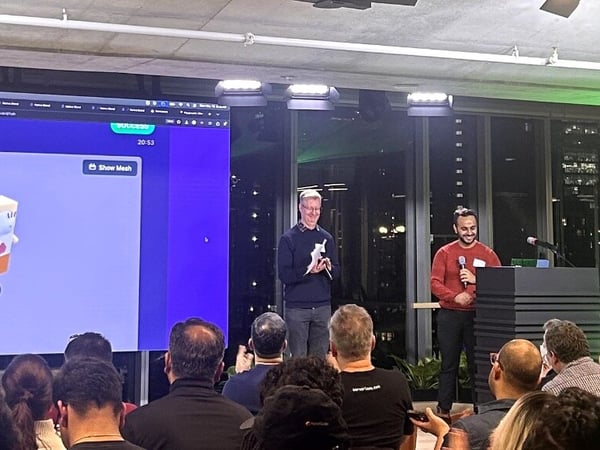 A photograph captures an indoor presentation event where two male speakers stand on stage, one holding a microphone, in front of a large screen displaying a 3D model of Spongebob Squarepants and generation status information.