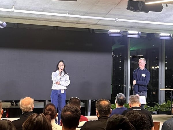 A photograph captures a presentation event where a young woman speaks into a microphone on a stage in front of a large, dark screen, while a man stands to her right; the foreground shows the backs of numerous audience members' heads.