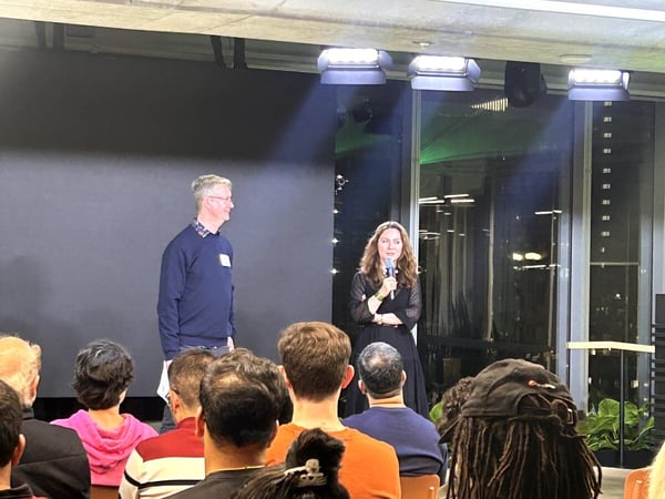 A photograph taken at an evening event shows a woman holding a microphone and a man standing on a stage area in front of a dark screen and large windows overlooking a city skyline at night, addressing an audience whose backs are visible in the foreground.