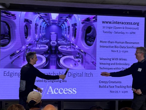 The image captures an indoor presentation event where two men are standing in front of a large projection screen displaying event schedules and a surreal, purple-toned image of a mirrored hallway filled with toilets and sinks.