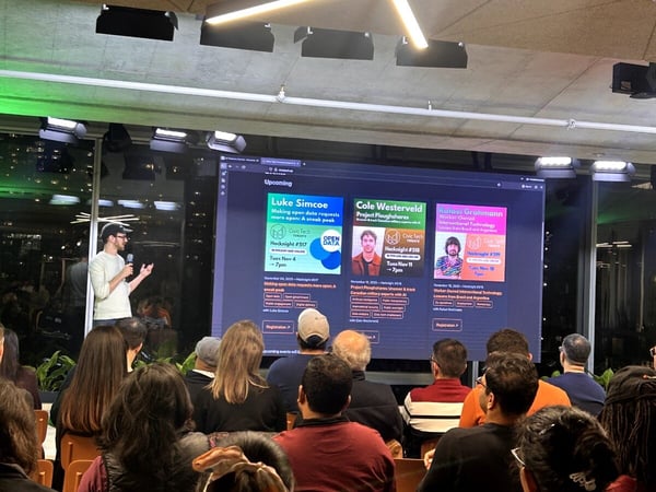 A photograph captures a speaker presenting at an evening tech event, standing beside a large screen displaying information about upcoming 'Hacknight' sessions to a seated audience.