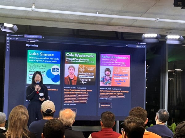 A photo capturing an indoor event where a woman is giving a presentation in front of a large screen displaying upcoming Civic Tech Hacknight events, while an audience watches from the foreground.
