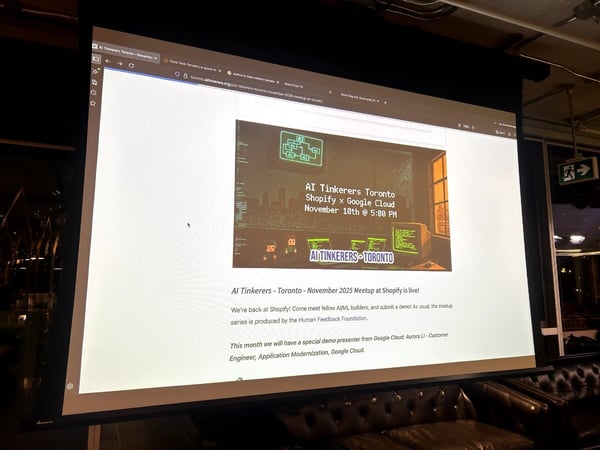 A photograph captures a large projection screen displaying a website for the 'AI Tinkerers - Toronto - November 2025 Meetup at Shopify' event, set above dark, tufted leather seating in a dimly lit indoor venue.