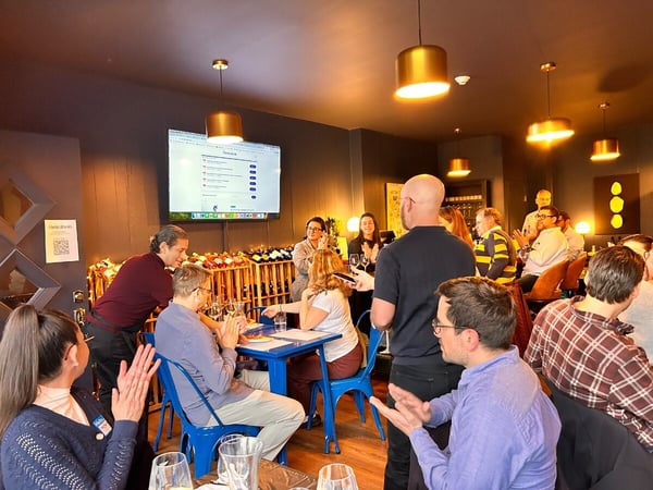 A group of people are gathered in a dimly lit restaurant or wine bar for a presentation, with several individuals clapping while looking at a screen displaying a website about Paris 2024.