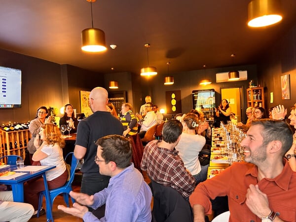 A group of people are gathered in a dimly lit restaurant or bar, many of whom are clapping while looking towards a screen or speaker.