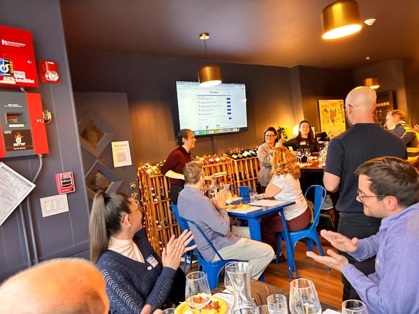 A group of people are gathered in a wine bar for a presentation or social event, with some seated at blue tables and others standing near a screen displaying a website.