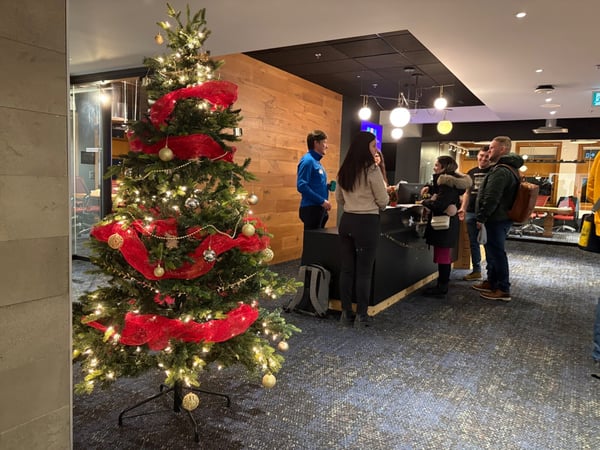 A photograph taken indoors in a modern lobby or reception area, featuring a brightly lit Christmas tree decorated with red ribbon and ornaments on the left, and a group of people gathered around a dark counter on the right.