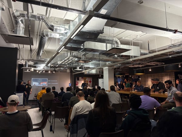 A photograph capturing an indoor presentation or event in a modern space with exposed ductwork, showing a speaker addressing a large audience seated in rows and gathered around a counter.