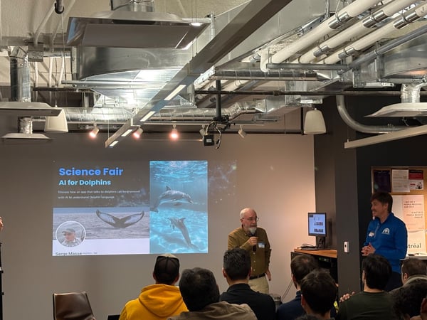 A photograph capturing an indoor technical presentation where a speaker, holding a microphone, addresses an audience in front of a screen displaying a slide about AI for Dolphins.