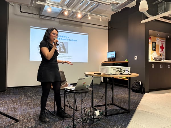 A woman wearing a black dress is giving a technical presentation indoors, holding a microphone while standing next to a table holding a projector and a laptop.