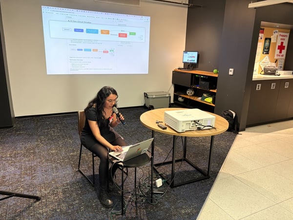A woman sits on a chair, speaking into a microphone while operating a laptop during a technical presentation in a dimly lit room. A flowchart titled "AI Ops Circuit Breaker" is projected onto a large screen behind her.